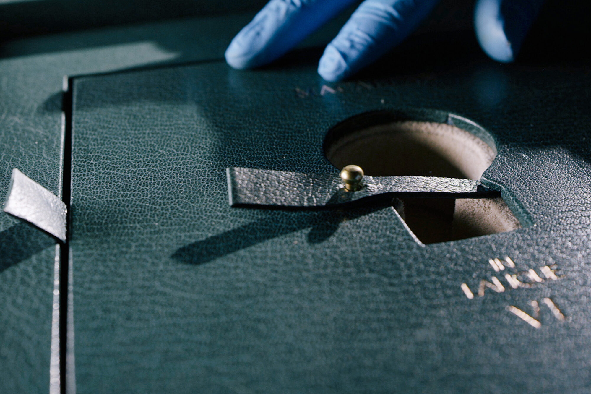 A gloved hand handles a green textured leather box with a circular cutout and a strap fastened by a gold button; some gold embossed letters are partially visible on the surface, resembling The Macallan case study packaging.
