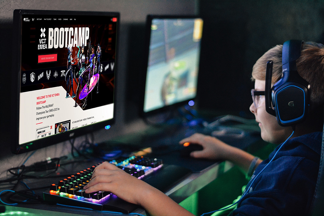 A young boy wearing headphones sits at a desk, playing a video game on a computer with a colorful backlit keyboard. The screen displays a Riot Games VCT gaming bootcamp website.