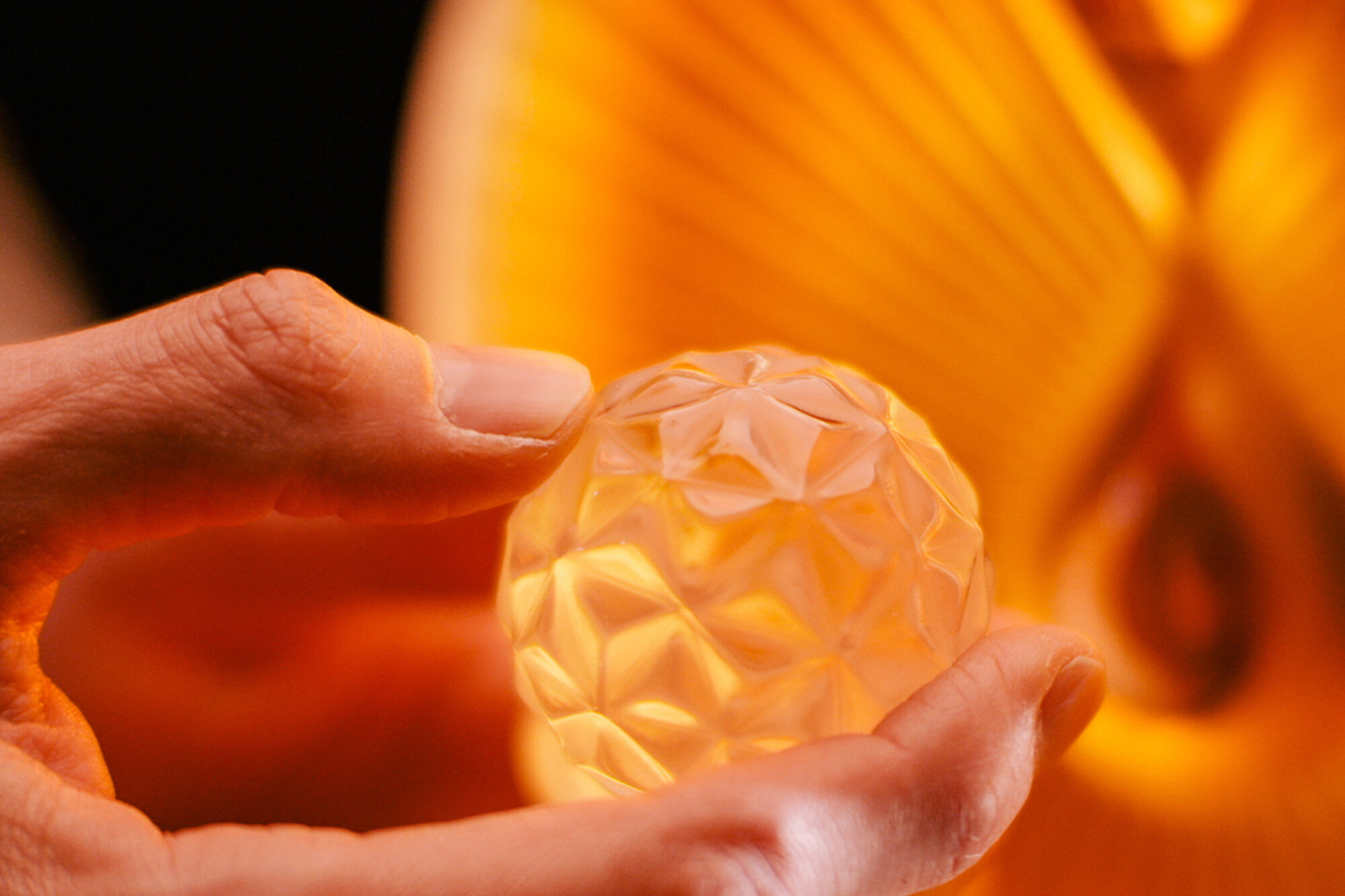 A hand gently holds a small, faceted, translucent sphere that glows with warm orange light, reminiscent of The Macallan case study, with an abstract golden background enhancing the luminous effect.