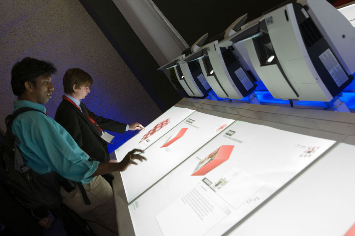 Two people interact with a large, illuminated touchscreen display featuring digital content and images, while several Hitachi Inspire Life desktop computers are positioned on a ledge above the screen.