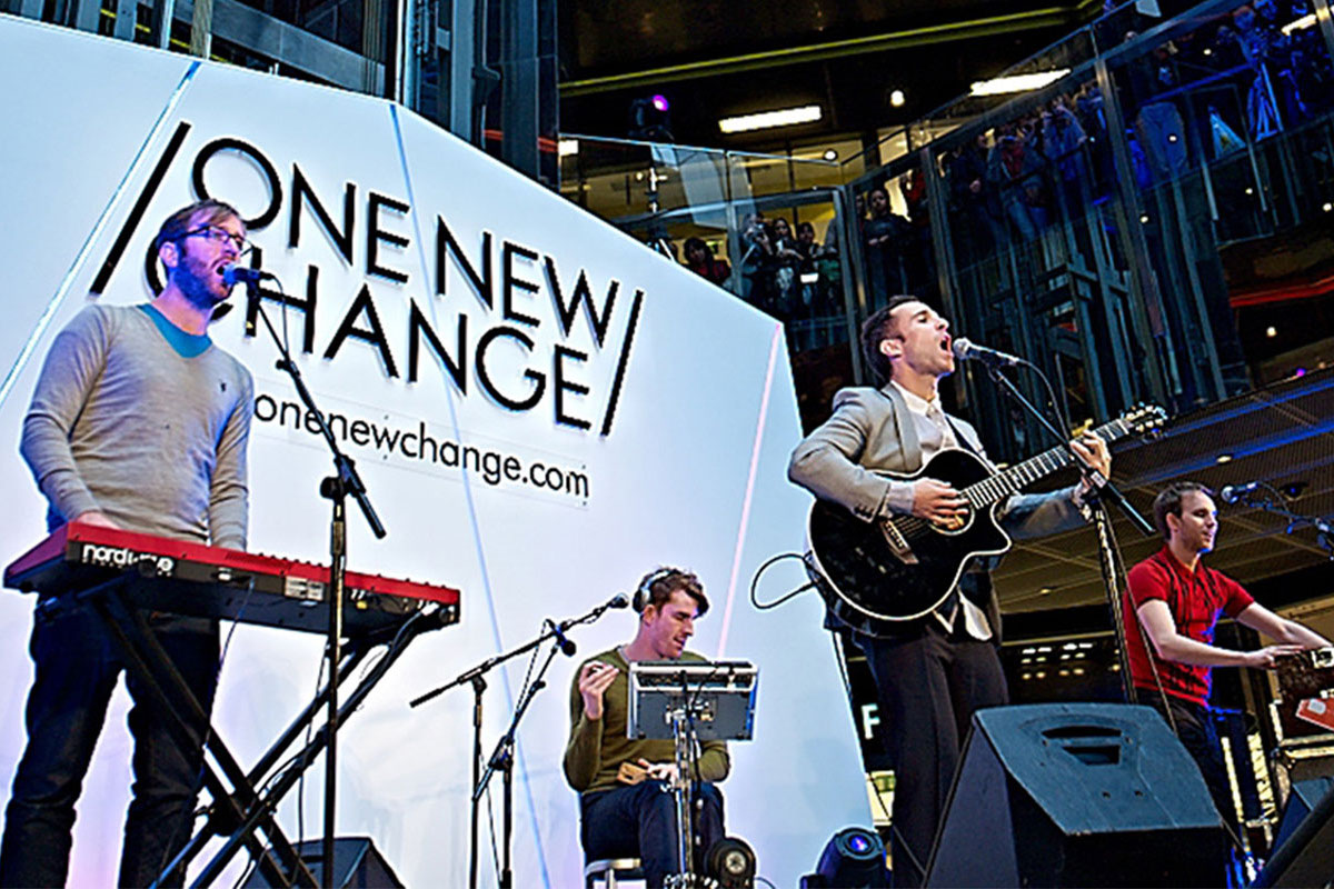 A four-member band performs live on a brightly lit stage labeled “One New Change,” part of a Landsec case study. One plays keyboard, another drums, the third sings and plays guitar, while the fourth also plays keyboard as an audience watches from multiple levels.