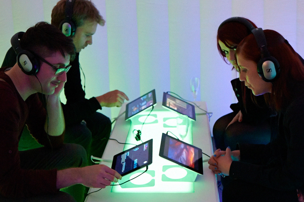 Four people wearing headphones sit around a glowing table, each interacting with a touchscreen tablet. The scene, part of a G-Tech case study, is set in a dimly lit room illuminated by green and blue lighting.