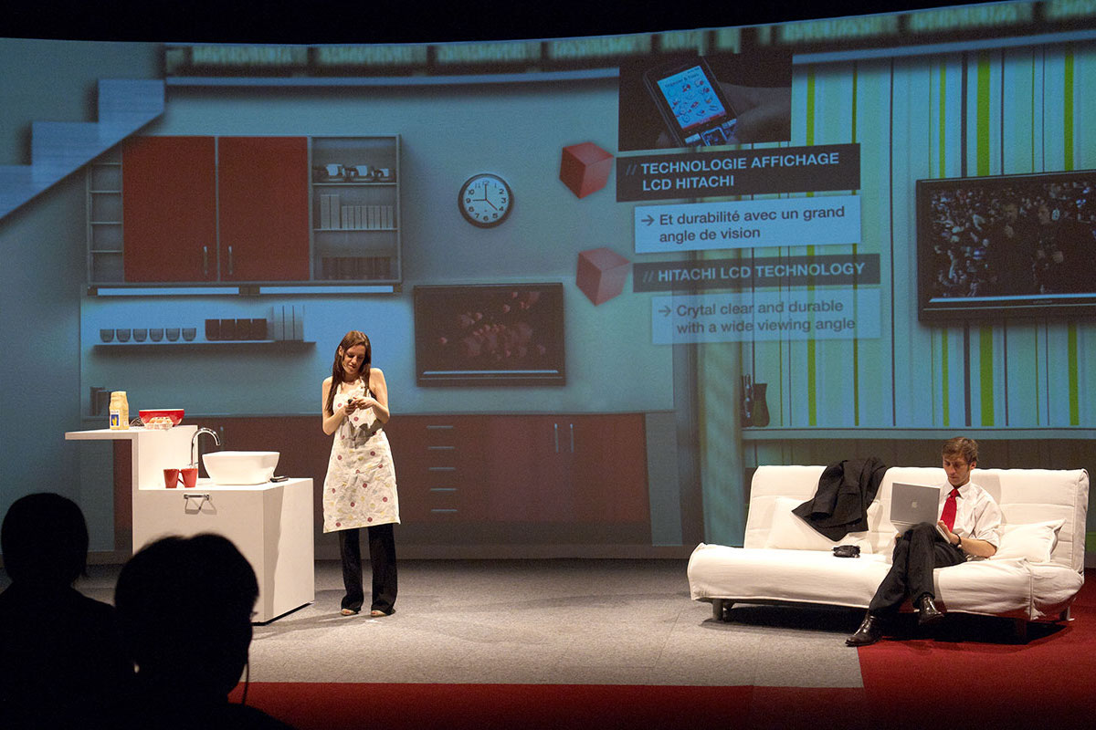 A woman in an apron stands on a stage beside a sink, while a man sits on a white couch with a laptop. Behind them, a large screen showcases Hitachi Inspire Life kitchen graphics and technology information in French and English.