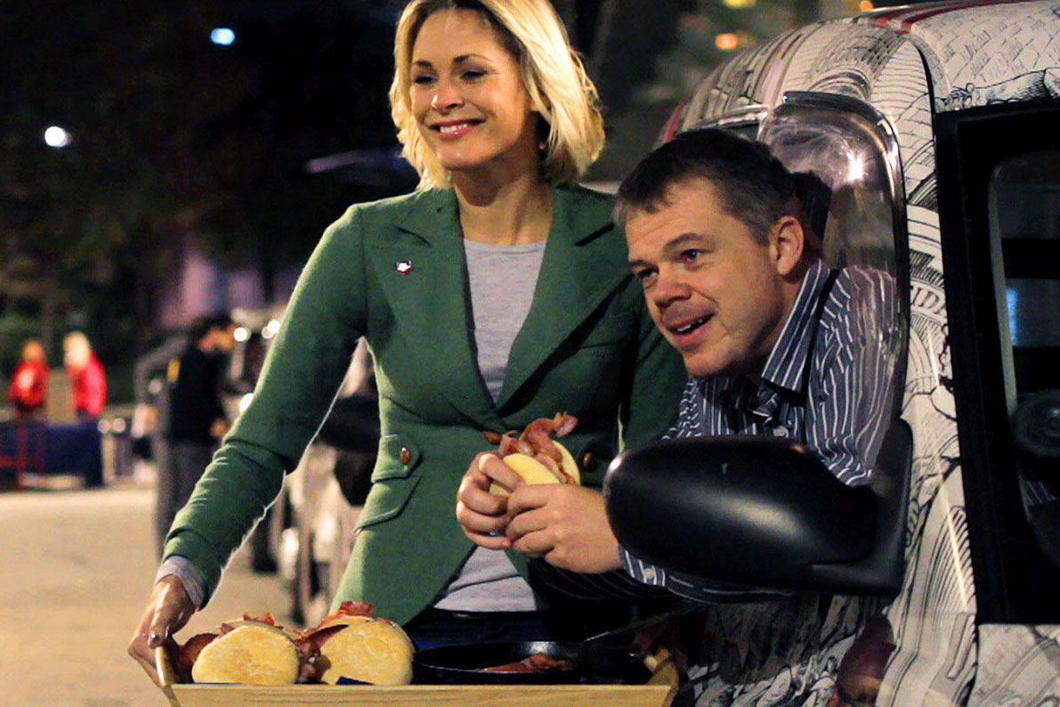 A woman stands next to a smiling man leaning out of a car window. They are both holding sandwiches and serving food from a tray with more sandwiches on it, outdoors at night, as part of the Landsec case study.