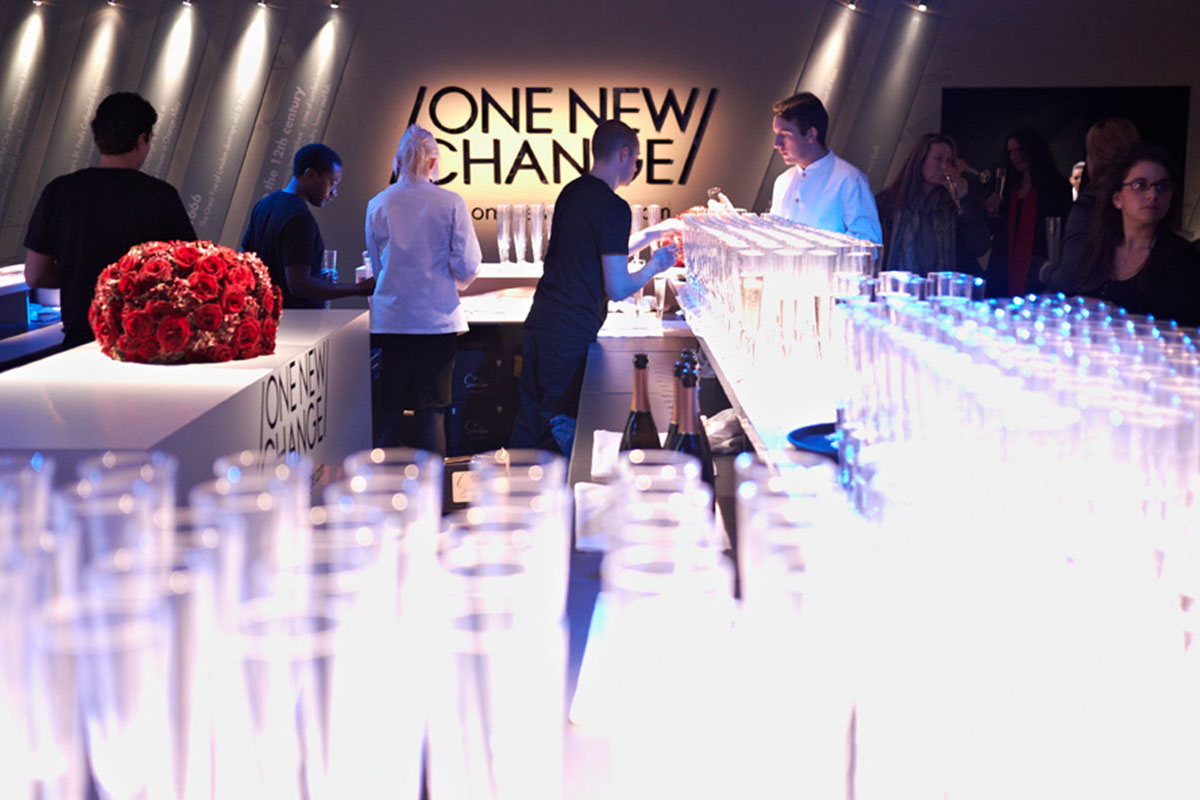 A modern bar with glowing blue and white lighting, filled with empty glasses. Bartenders serve drinks to guests, and a centerpiece of red roses sits on the counter—a striking scene from the Landsec case study at One New Change.