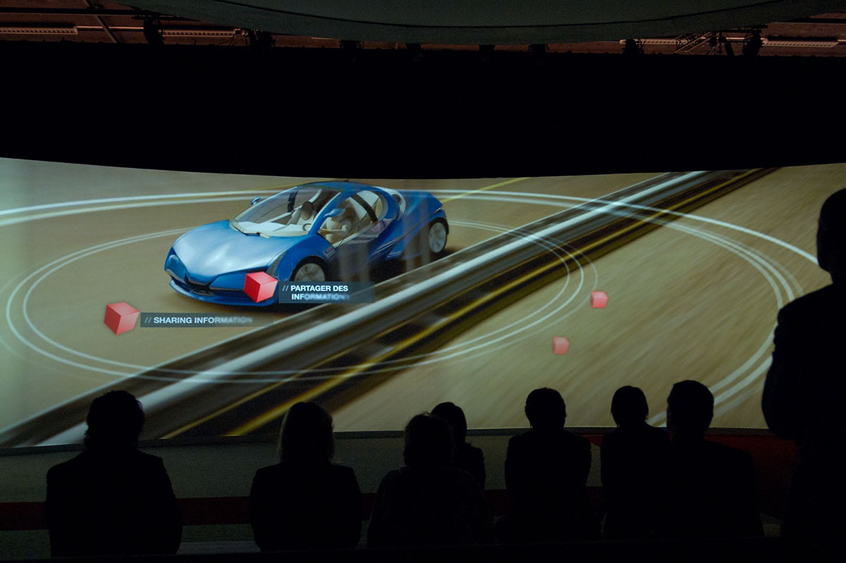 A group of people sit in shadow watching a large screen displaying a blue futuristic car with digital graphics and information overlays, reflecting the innovative spirit of Hitachi Inspire Life, in a dark room.
