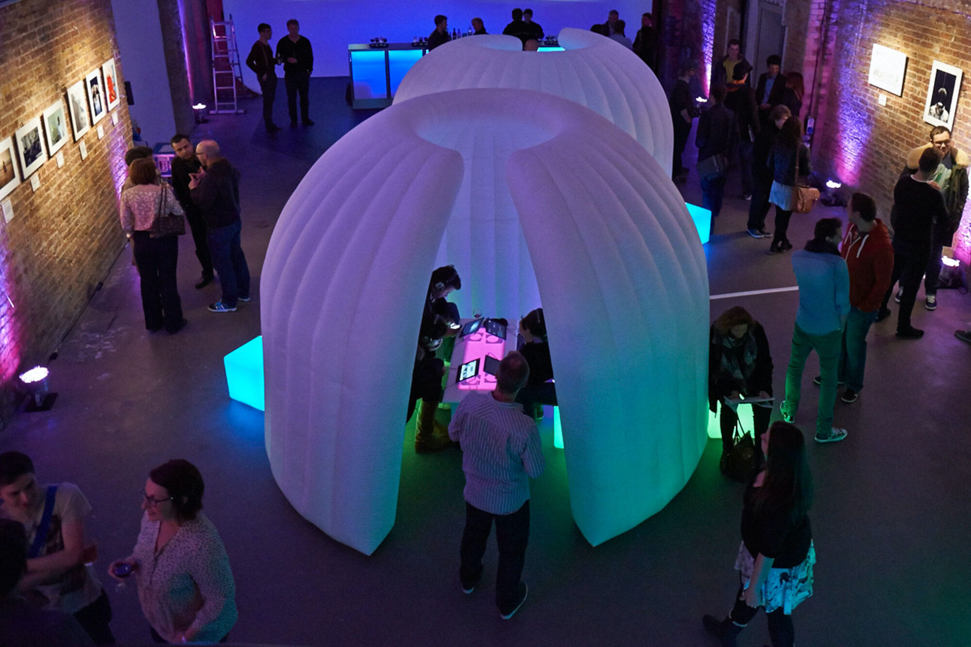 People mingle at an indoor event with brick walls and colorful lighting. A large white, dome-shaped inflatable structure, featured in the G-Tech case study, is in the center, with attendees interacting around and inside it. Framed photos hang on the walls.