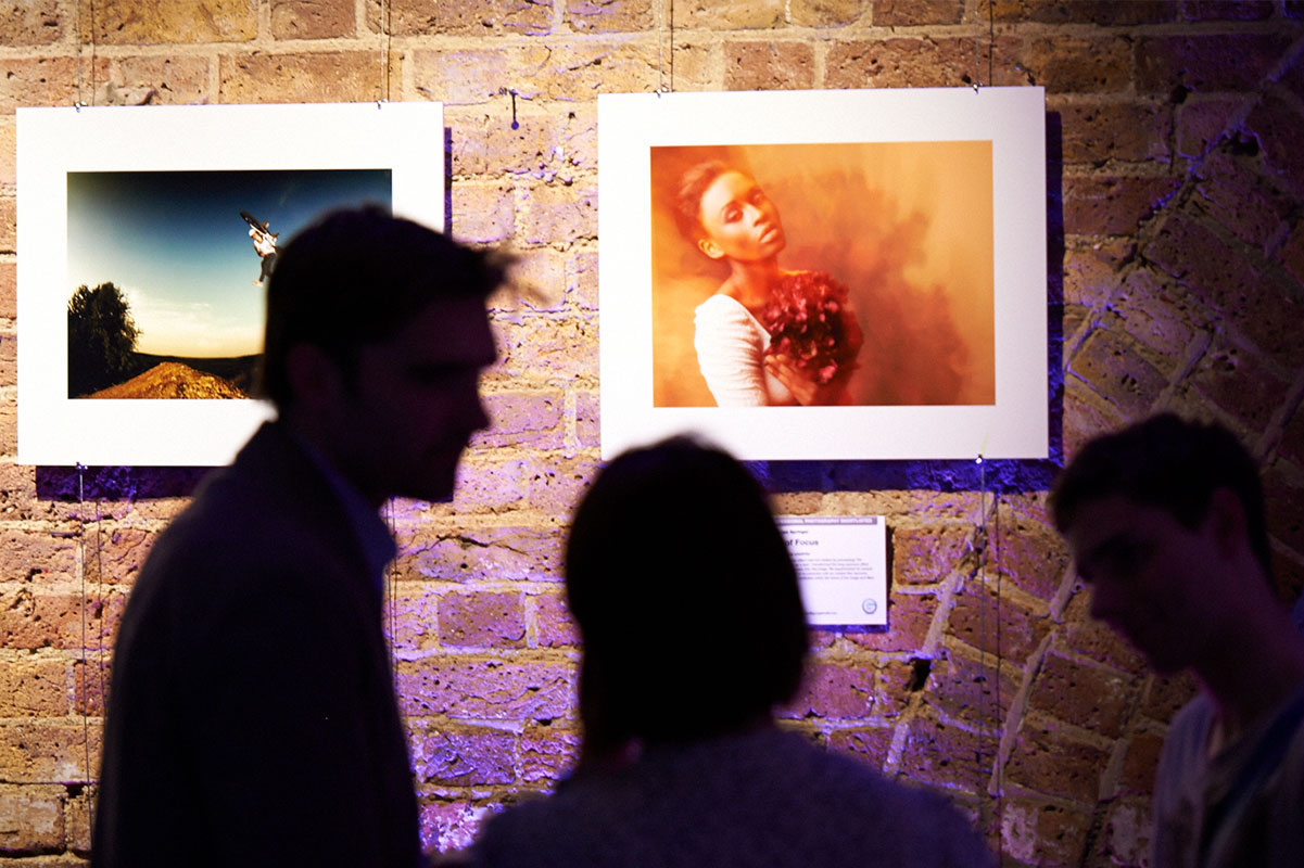 Three people in silhouette view framed photographs displayed on a brick wall in this G-Tech case study; one photo shows a person holding flowers, while another shows someone standing on a dirt mound, partially illuminated by blue light.