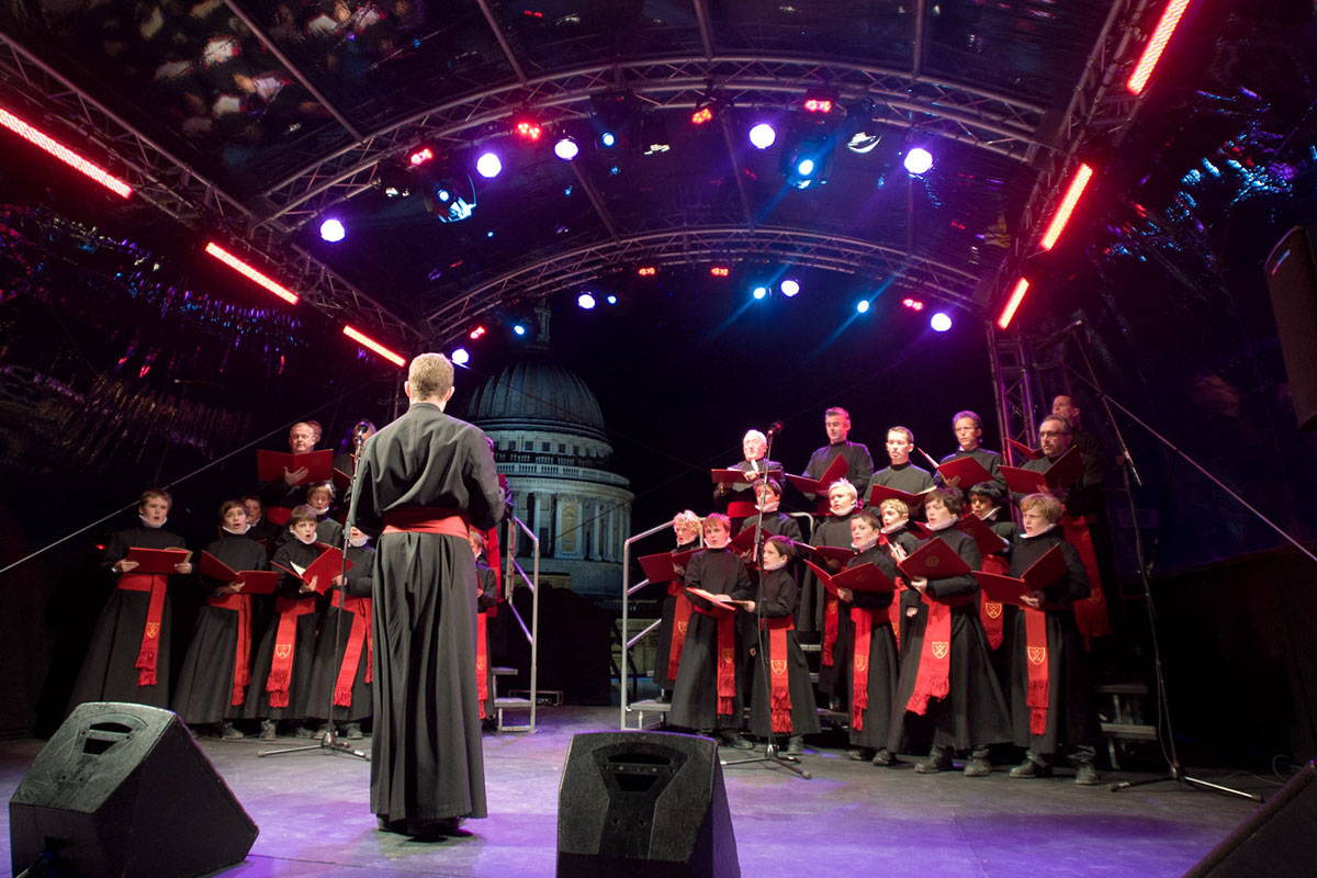 A choir dressed in black robes with red sashes sings on an outdoor stage under colorful lights, led by a conductor, with a domed building in the background—a vibrant scene reminiscent of a Landsec case study in community engagement.