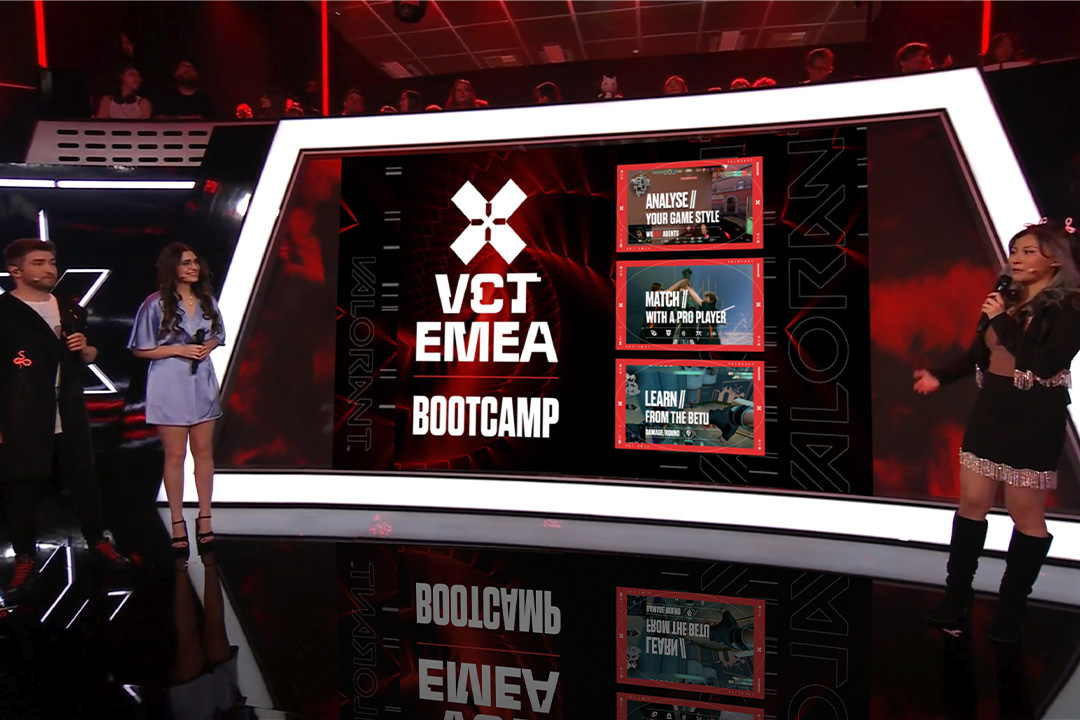 Three presenters stand on stage next to a large screen displaying Riot Games VCT EMEA Bootcamp and three event highlights. The stage glows with red and black lighting as an audience watches from the background.