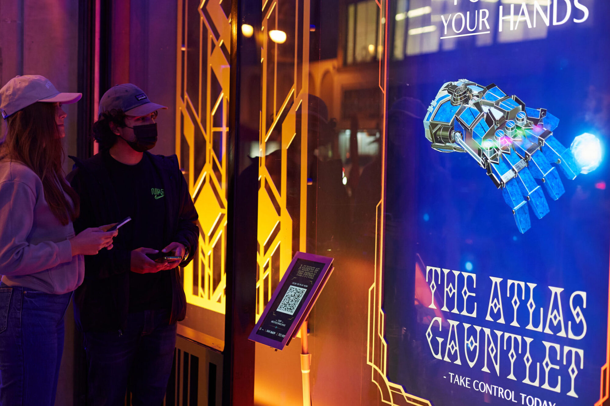 Two people stand by a brightly lit interactive display featuring a large robotic hand and text reading The Atlas Gauntlet - Take Control Today. One holds a phone, reminiscent of Netflix Arcanes tech-inspired visuals, as they both look at the screen.