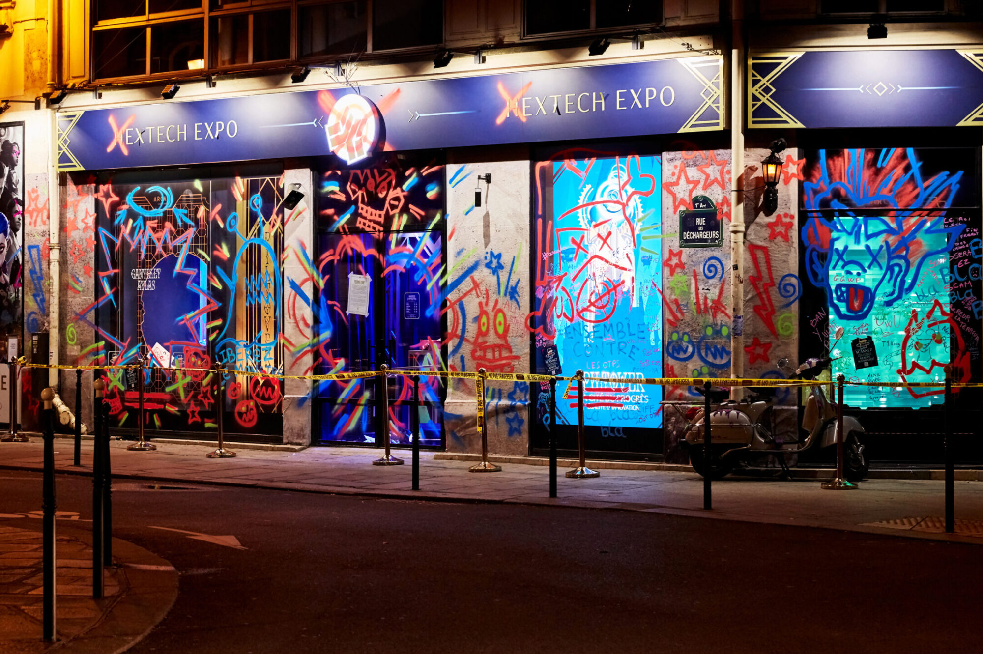 A storefront at night with windows covered in colorful graffiti art inspired by Netflix Arcane, featuring faces, shapes, and bright neon colors. The sign above reads XETECH EXPO and yellow caution tape lines the sidewalk in front.