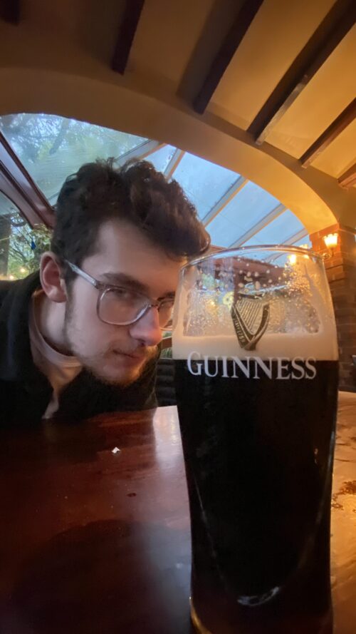 A young man with glasses, part of the marketing team, looks closely at a pint of Guinness on a wooden table in a cozy pub with large windows and a warm atmosphere.