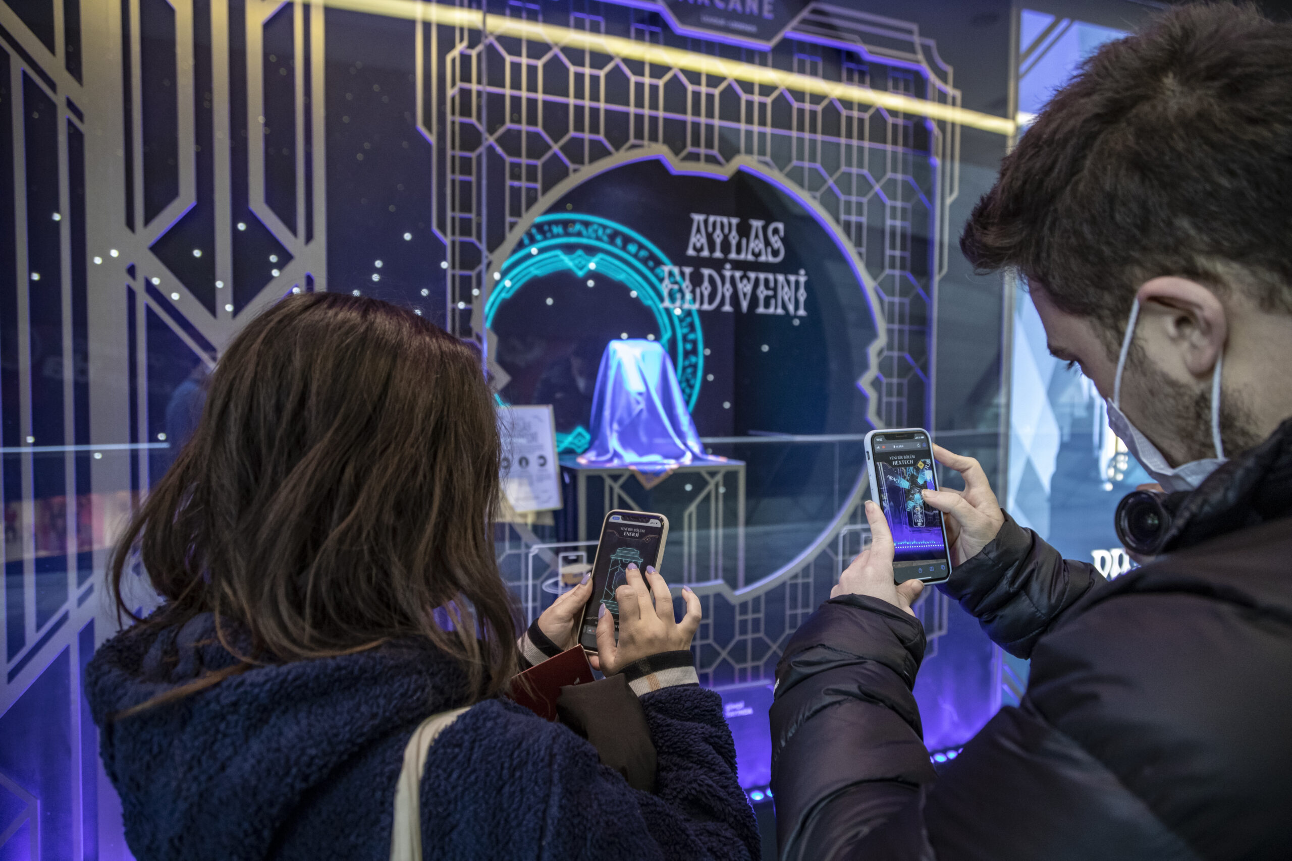 Two people snap photos with their phones of a display window featuring a covered object and a decorative sign reading Atlas Advent. The art deco scene glows with blue and purple lights, evoking the vibrant style of Netflix Arcane.