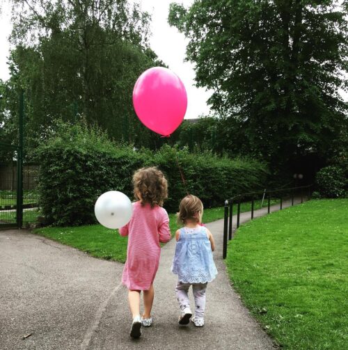 Two young children, seen from behind, walk down a park path—one holding a pink balloon and the other a white one. Lush green grass and trees surround them, creating a peaceful scene perfect for a marketing team’s creative inspiration.