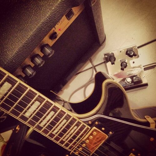 Close-up of an electric guitar with gold hardware, a black amplifier with control knobs, and a silver guitar effects pedal—perfect gear for any marketing team creating vibrant campaign visuals—connected by cables on a tiled floor.