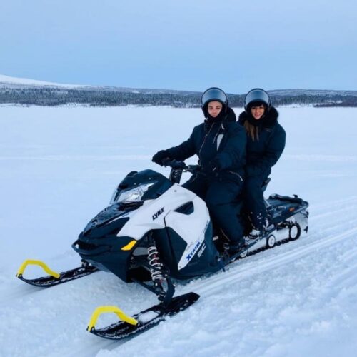 Two people wearing helmets and winter jackets sit together on a snowmobile in a snowy, open landscape with distant trees and a cloudy sky, capturing the spirit of experiential marketing in an adventure that builds unforgettable loyalty.