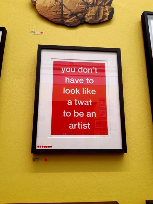 A framed artwork with a red background and white text reads, you don’t have to look like a twat to be an artist, displayed on a yellow wall—perfect inspiration for any marketing team in an art gallery.