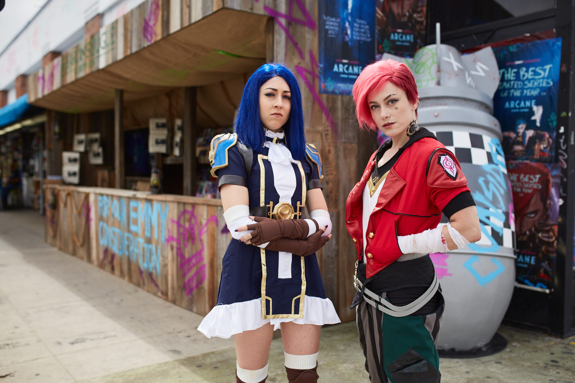 Two people in detailed Arcane FYC Emmys cosplay pose confidently on a city sidewalk in front of a graffiti-covered building and a large prop. One wears a navy and white outfit, the other a red and black ensemble with short pink hair.
