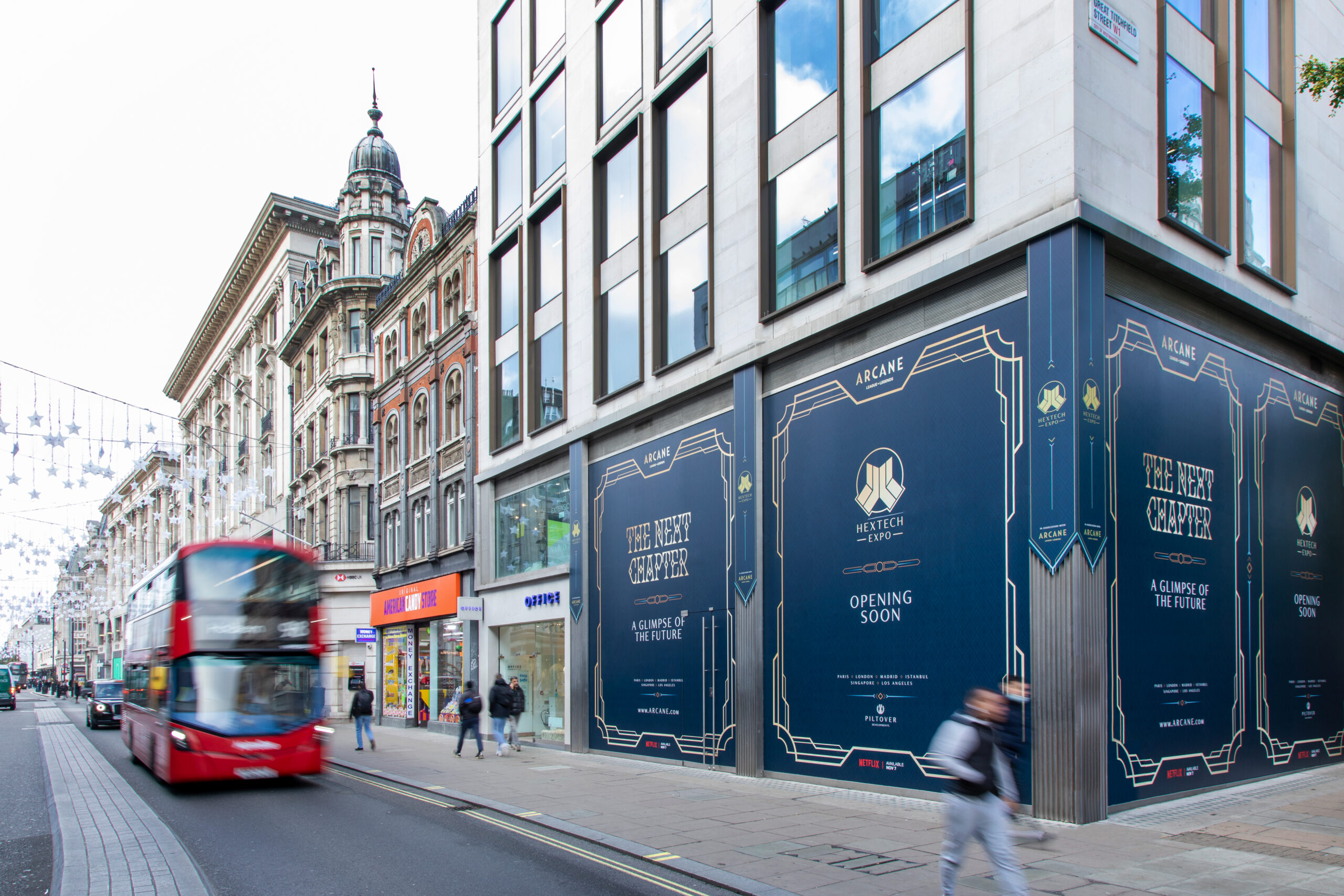 A red double-decker bus passes by a city street lined with shops and historic buildings; one corner building is covered in blue and gold MetTech signs announcing The Next Chapter, Opening Soon, and a Netflix Arcane poster. Pedestrians walk nearby.