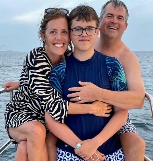 A smiling woman, teenage boy, and man—like a cheerful marketing team—pose together on a boat with the ocean in the background. The woman wears a zebra-print cover-up, the boy sports glasses and a blue swim shirt, and the man is shirtless in patterned trunks.