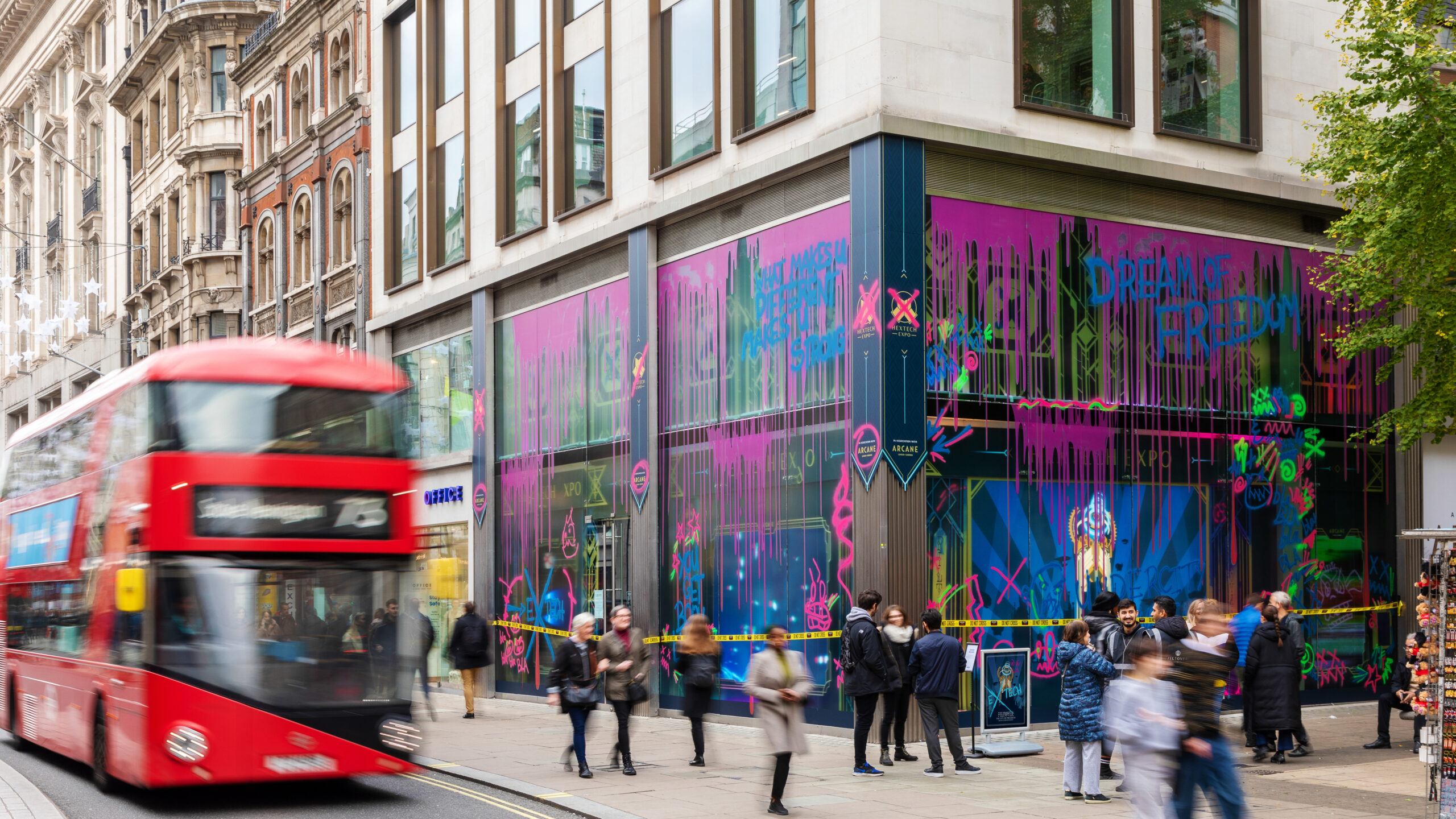 A red double-decker bus passes by a busy London street corner, where people walk in front of a building decorated with bright, Netflix Arcane-inspired graffiti-style art on its windows.
