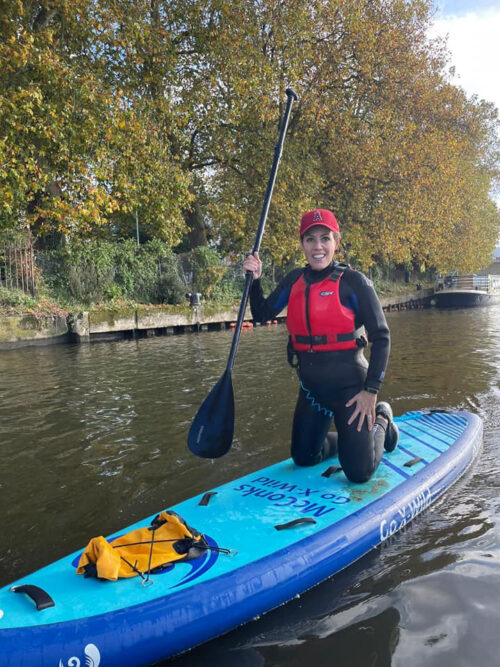 A person wearing a red life jacket, black wetsuit, and red cap is kneeling on a blue stand-up paddleboard, smiling as if celebrating with their marketing team on a calm river with trees and a canal bank in the background.