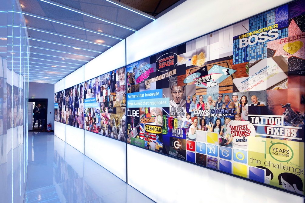 A brightly-lit hallway with glowing walls, one side covered in a collage of colorful TV show logos and images—including titles like Undercover Boss, Tattoo Fixers, and 10 Years Younger—creates the vibe of an All3Media case study come to life.