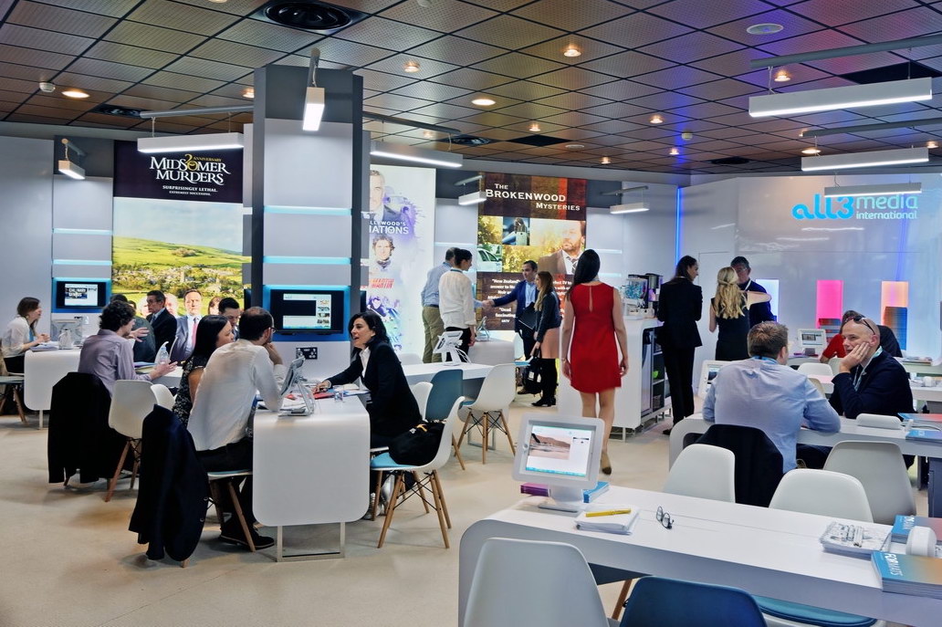 People in business attire sit and stand around tables at a busy convention or trade show, with All3Media case study posters for TV shows like Midsomer Murders and Brokenwood Mysteries displayed in the background.