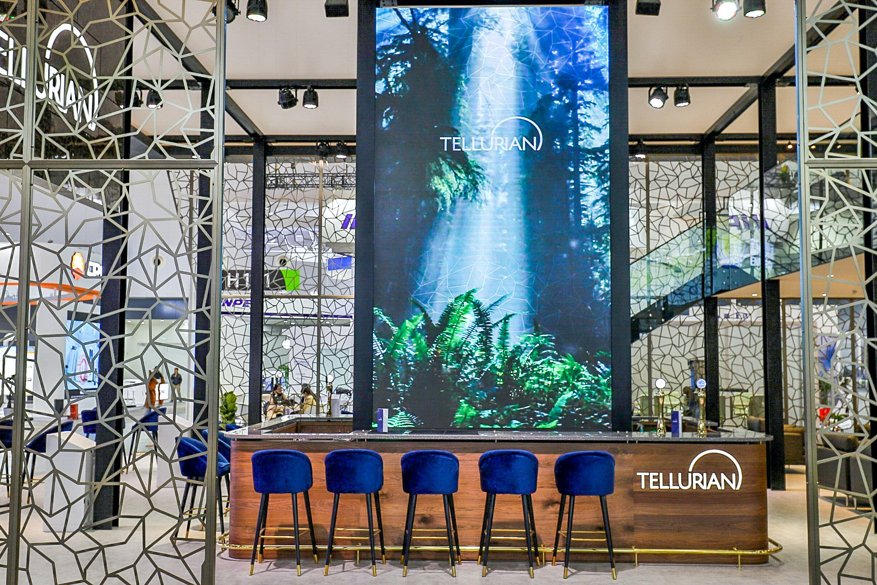 A modern exhibition booth for Tellurian features a wooden bar with five blue velvet stools and a large digital screen displaying a forest scene, inspired by the Tellurian case study. Geometric patterned panels stylishly frame the booth.