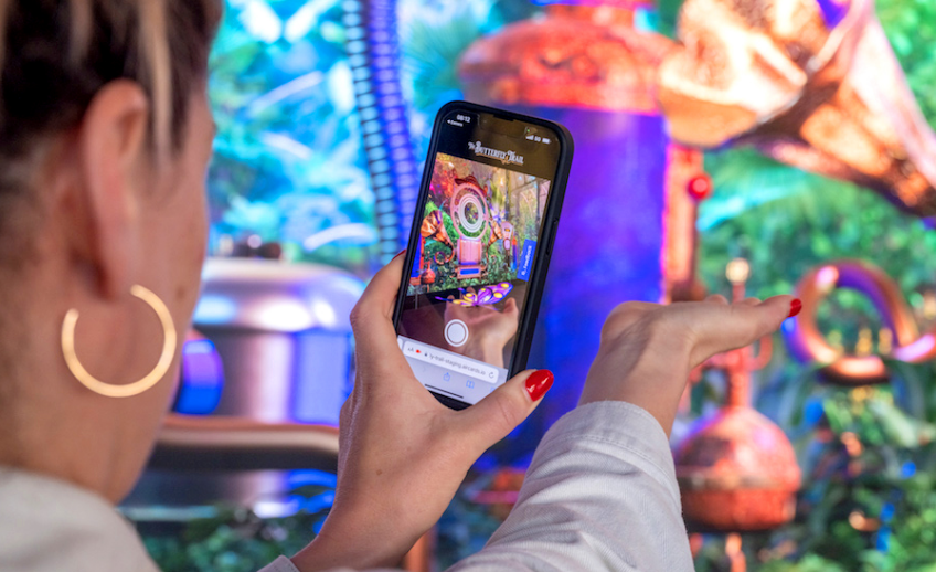 A person with red nails and hoop earrings holds up a smartphone, capturing a photo of a colorful, whimsical display in an innovation lab filled with various brass objects and lush greenery in the background.