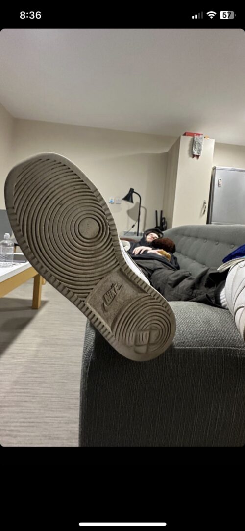 A person is lying on a couch with their foot extended toward the camera, showing the sole of a Nike shoe—a relaxed setting that could inspire any marketing team. The living room has a table, lamp, fridge, and another person’s legs visible.