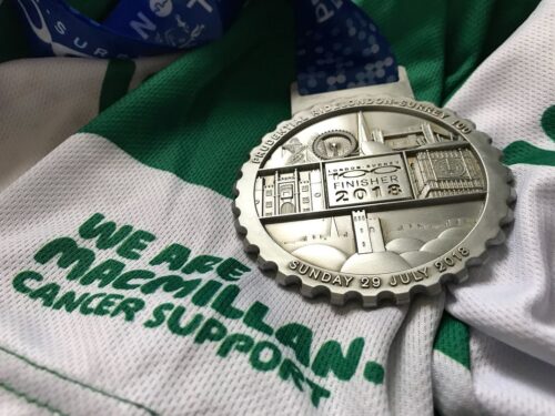 A silver finisher medal dated Sunday 29 July 2018 rests on a green and white We Are Macmillan Cancer Support shirt, a testament to the marketing teams dedication, with a blue lanyard partially visible in the background.