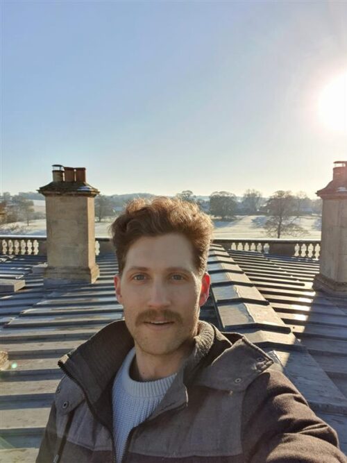 A man with light brown hair and a mustache, part of a marketing team, takes a selfie on a sunny rooftop, with stone chimneys and a scenic, misty landscape in the background.
