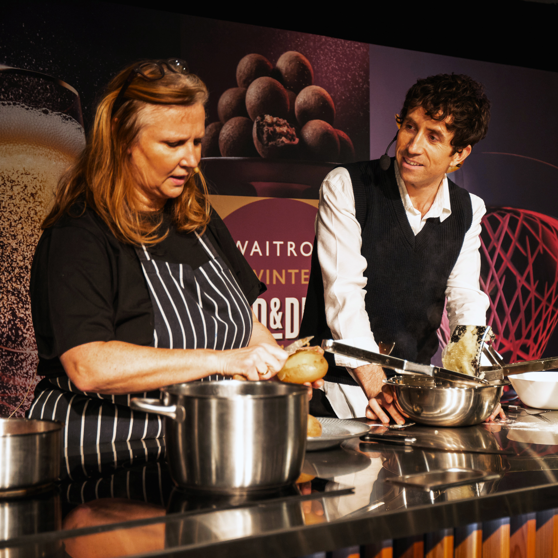 Two people stand behind a kitchen counter cooking, preparing dishes inspired by the Waitrose Winter Festival. The woman on the left peels a potato while the man in a vest smiles at her, surrounded by pots and kitchen utensils.