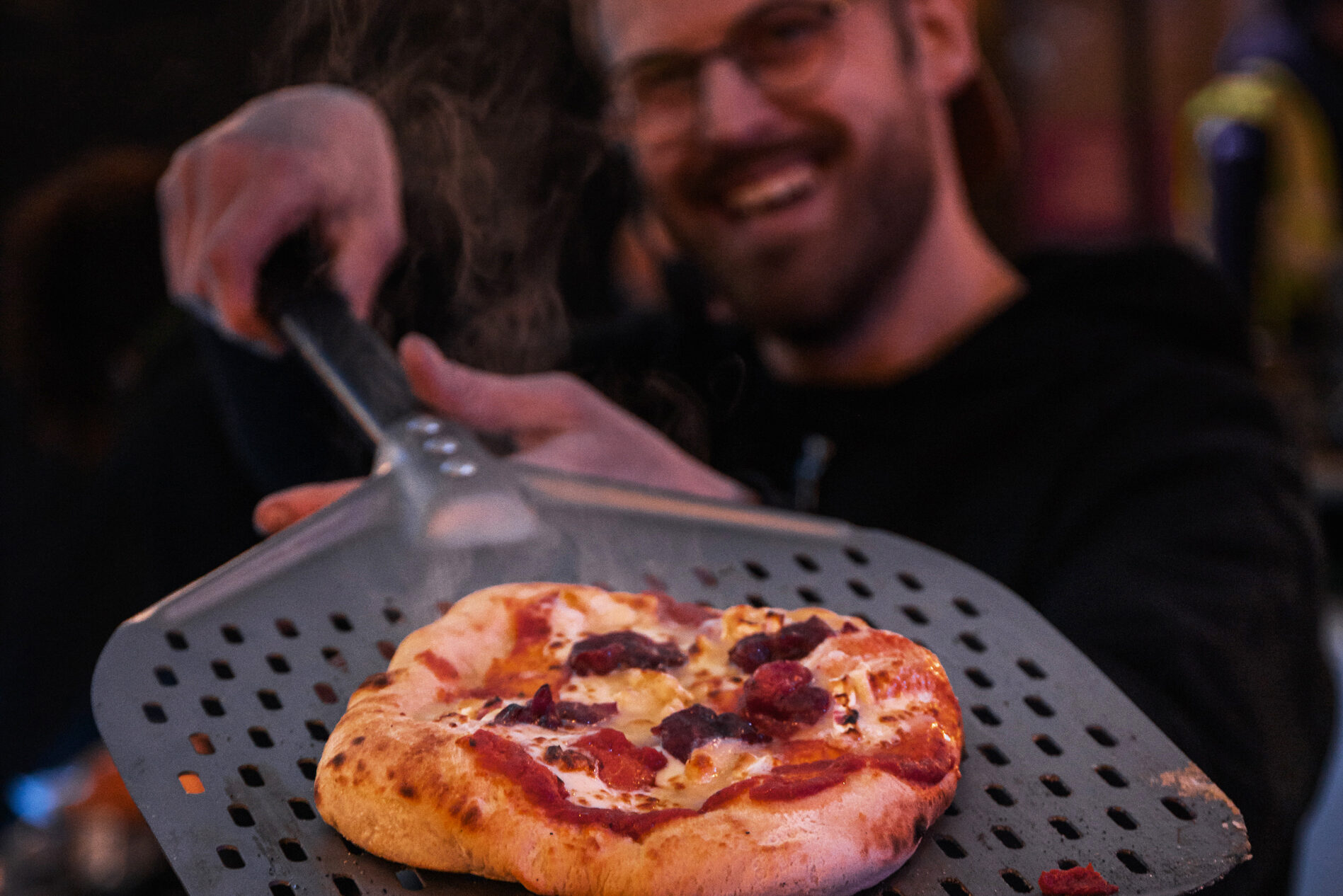 A smiling person holds a pizza peel with a freshly baked, steaming pizza topped with cheese and sausage, reminiscent of cozy treats enjoyed at the Waitrose Winter Festival, with the focus on the pizza in the foreground.