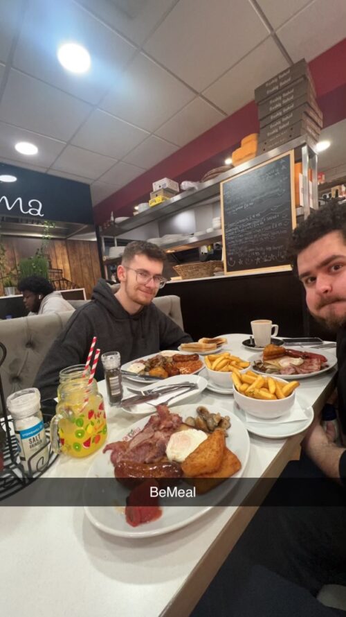 Two young men, perhaps from a marketing team, sit at a restaurant table filled with fries, toast, eggs, and bacon. One smiles at the camera while the other makes a playful face. The caption BeMeal appears at the bottom.