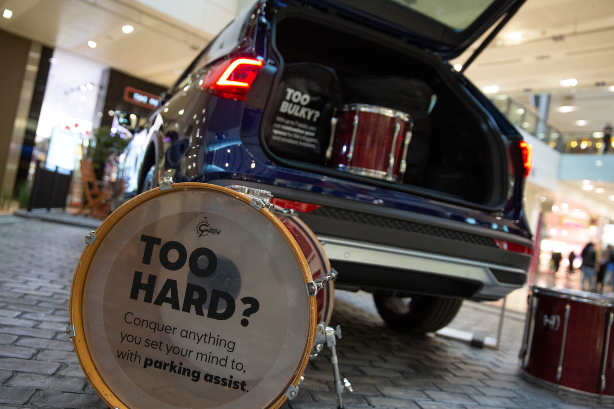A car with its trunk open is parked indoors, containing a drum set. A drum in the foreground displays: TOO HARD? Conquer anything you set your mind to, with parking assist—discover more in this SEAT case study.