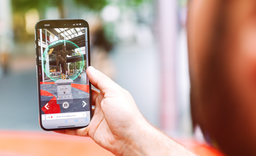 A person holds a smartphone displaying an augmented reality app, developed in an innovation lab, that overlays digital information about a statue or monument, with the real-world scene visible in the background.