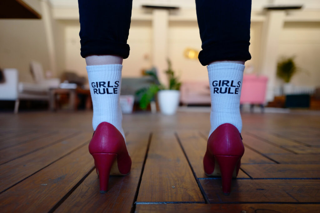 A person wearing white socks that say GIRLS RULE and red high heels stands on a wooden floor in a brightly lit room with modern, CSR and sustainability-inspired furniture in the background.