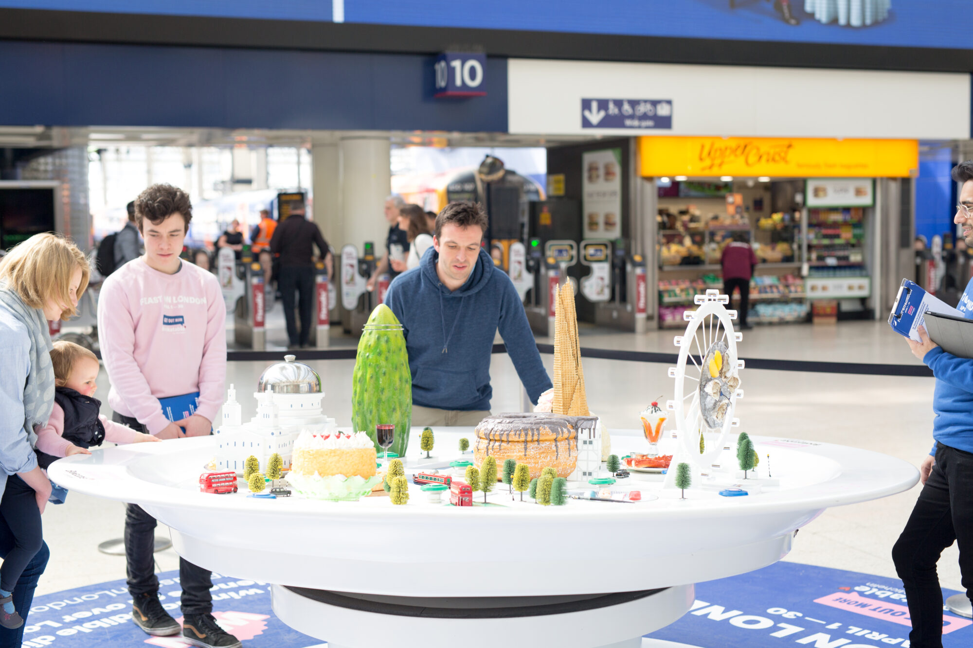 Several people gather around a large circular table featuring a colorful miniature city model, reminiscent of the collaborative planning seen in the Bookatable case study, with shops and signs visible in the busy indoor public space.