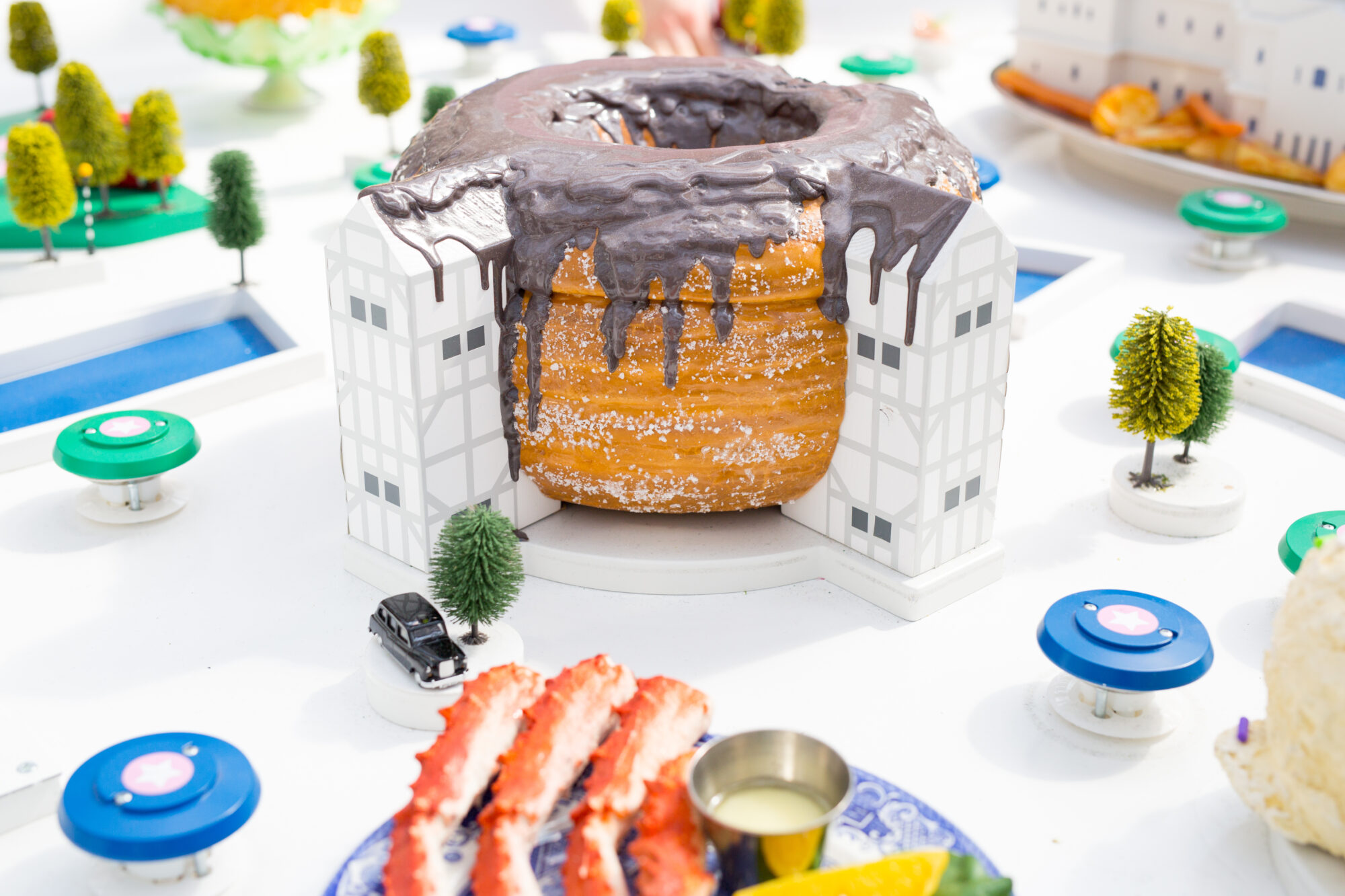 A large pastry topped with chocolate glaze is placed between two model white buildings, resembling an architectural landmark in a Bookatable case study. Miniature trees, toy cars, and colorful plates with food surround the scene.