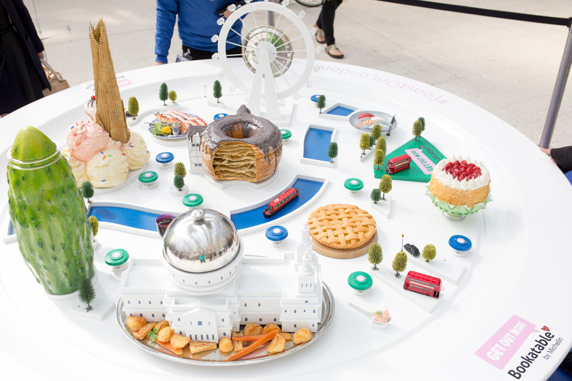 A circular table displays miniature London landmarks and famous foods, including a Ferris wheel, pie, pastries, and a large pickle—much like a scene from the Bookatable case study—arranged on a white surface with small trees and vehicles.