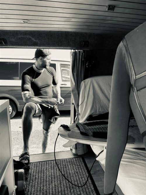 A man in a wetsuit stands by the open door of a van, stepping outside while holding gear—perhaps heading to meet his marketing team. Inside the van, a surfboard, wires, and part of a bed are visible in this black-and-white scene.