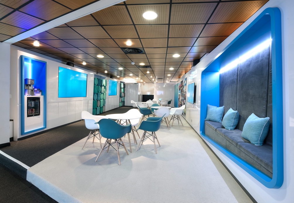 A modern office lounge with white tables, teal and white chairs, and built-in seating with blue accent lighting, designed as part of the All3Media case study. Wood panel ceilings and accent walls enhance its sleek, contemporary style.