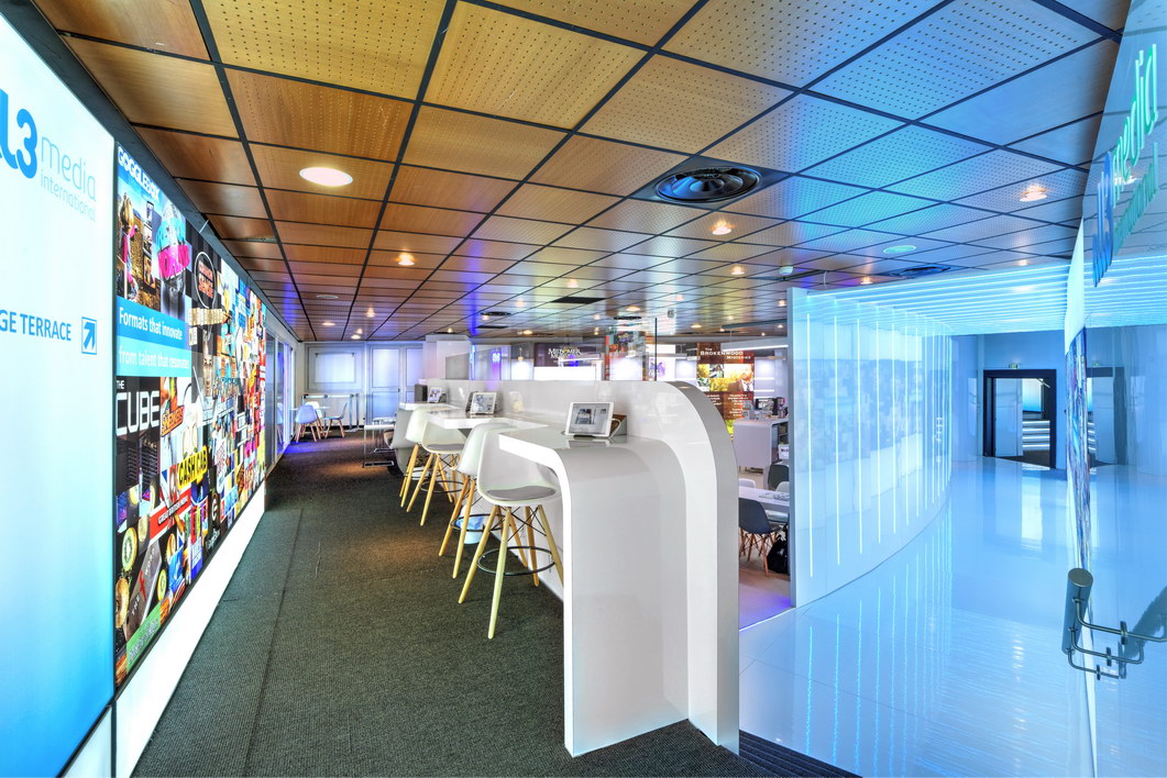A modern, brightly lit workspace with tall white desks and bar stools, digital screens displaying colorful images from the All3Media case study, and sleek blue walls and flooring in the background.