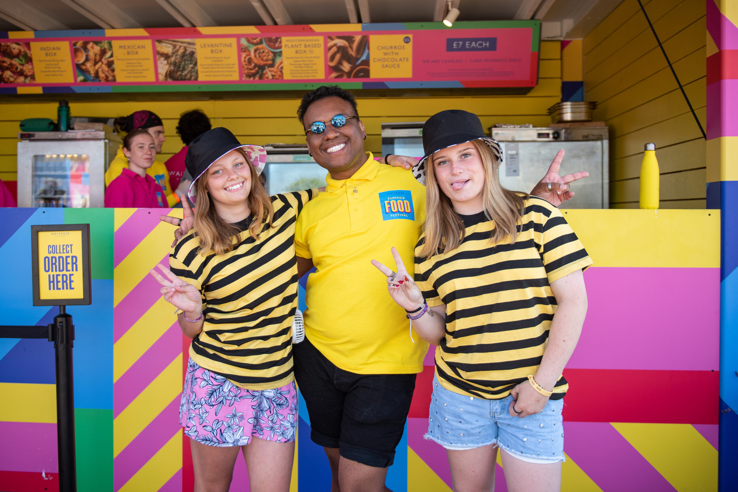 Three smiling people pose in front of a colorful food stand at Waitrose Summer Festivals. Two girls wear black-and-yellow striped shirts, black hats, and show peace signs. The staff member in the center sports a yellow “FOOD” polo and sunglasses.