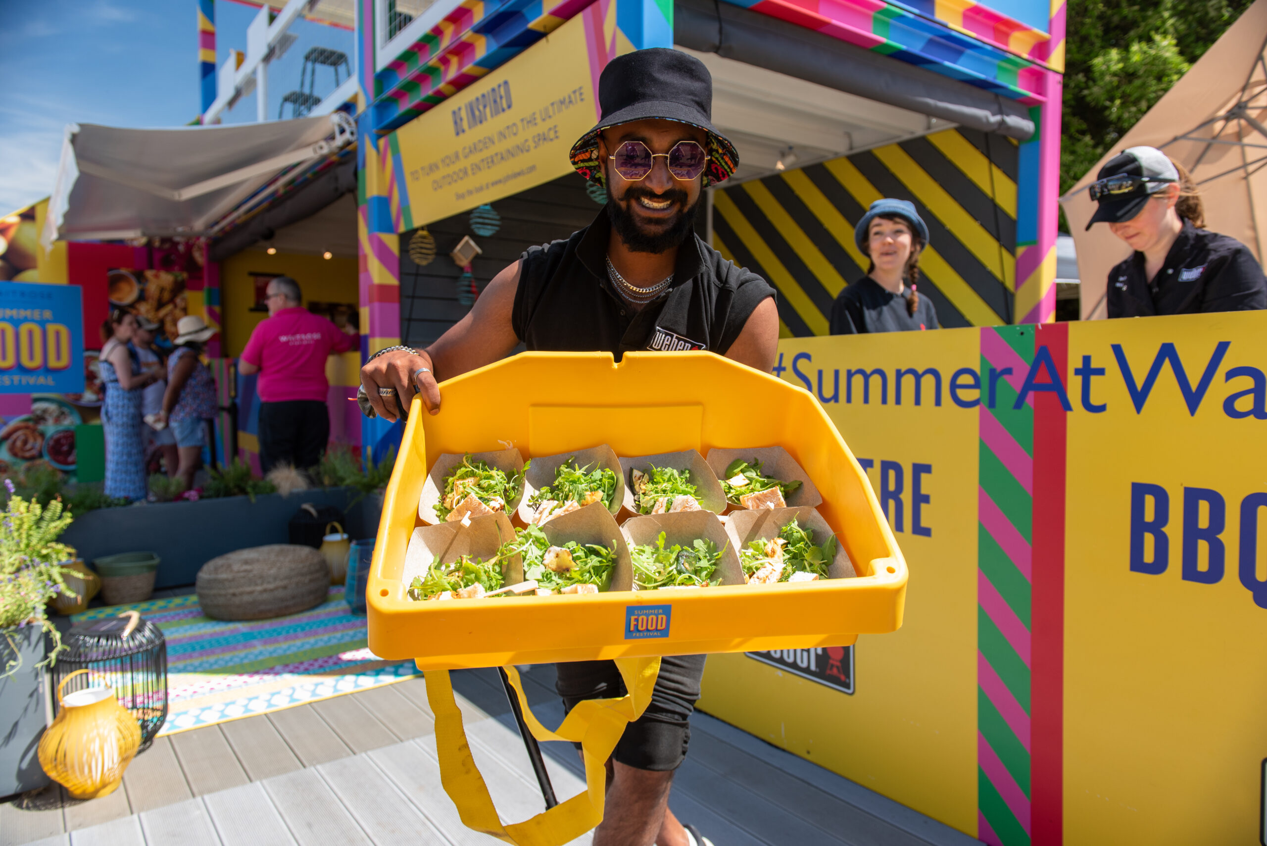 A smiling person in sunglasses and a hat holds a large yellow tray filled with small servings of food, standing at the vibrant Waitrose Summer Festivals with people and colorful signs in the background.
