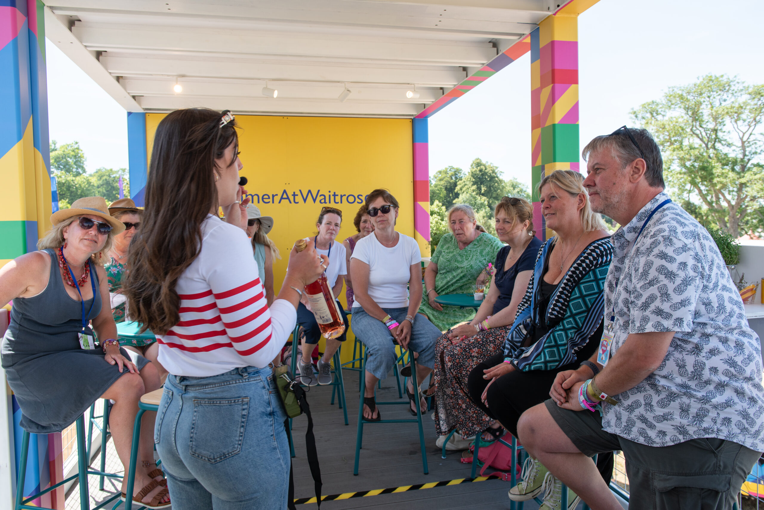 A woman addresses a seated group at an outdoor event, colorful Summer At Waitrose signage in the background. The group listens intently amid trees, enjoying one of the vibrant Waitrose Summer Festivals.