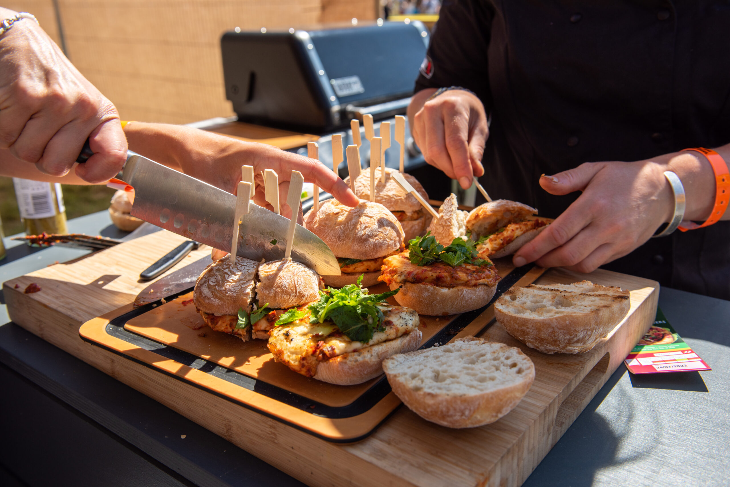Two people assemble chicken sandwiches with fresh greens on a wooden board outdoors, using knives and securing the buns with small wooden clips. Sunlight highlights the scene, evoking the relaxed spirit of Waitrose Summer Festivals.